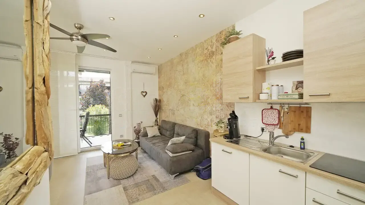 Wohnzimmer mit grauem Sofa, Einbauküche und Glastür mit Balkon